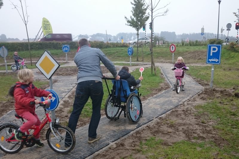 Kinderen met fietsjes en kin in rolstoel spelen samen op een verkeerspleintje met echte verkeersborden